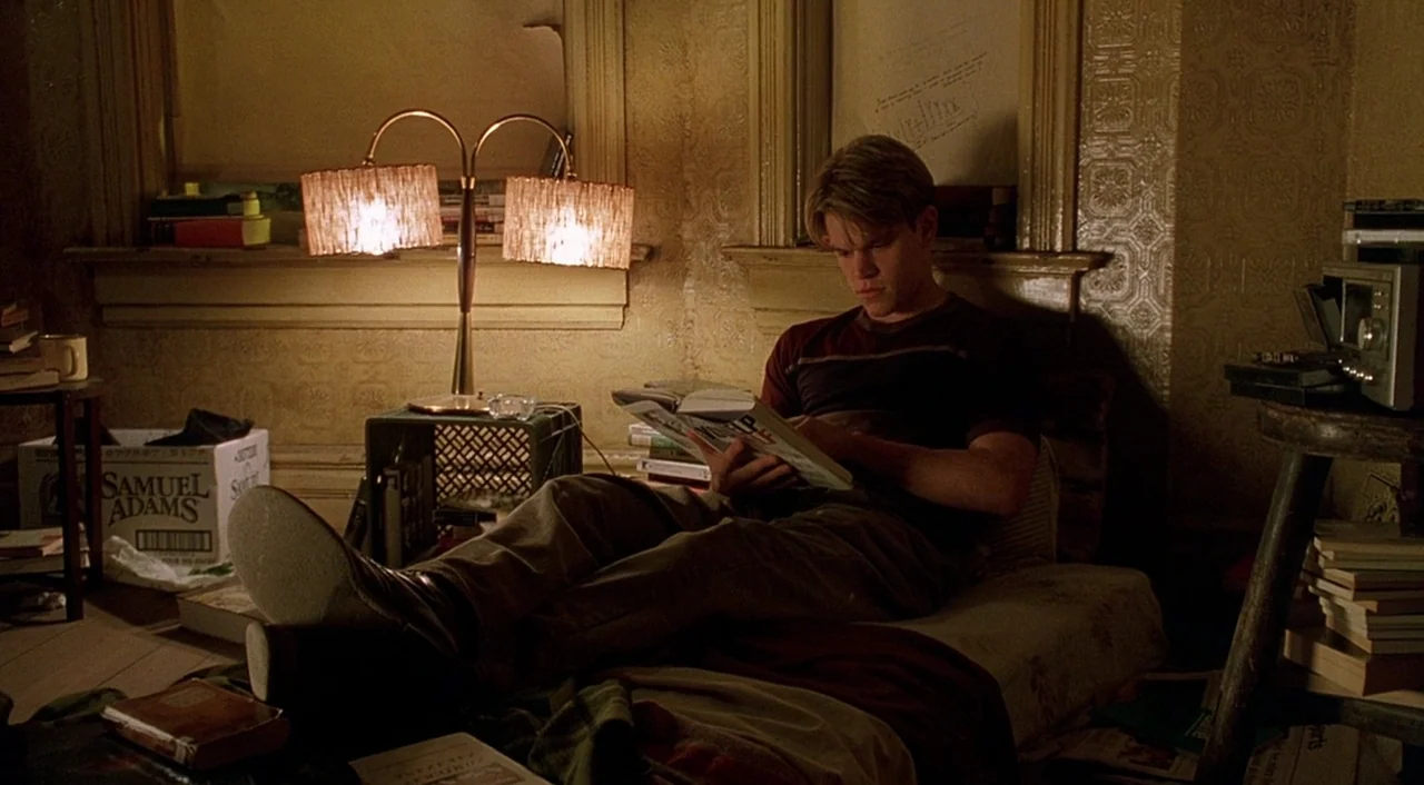 Good Will Hunting (1997) movie still: medium — A dimly lit, cramped room features patterned wallpaper and stacks of books. A young man reclines on a…