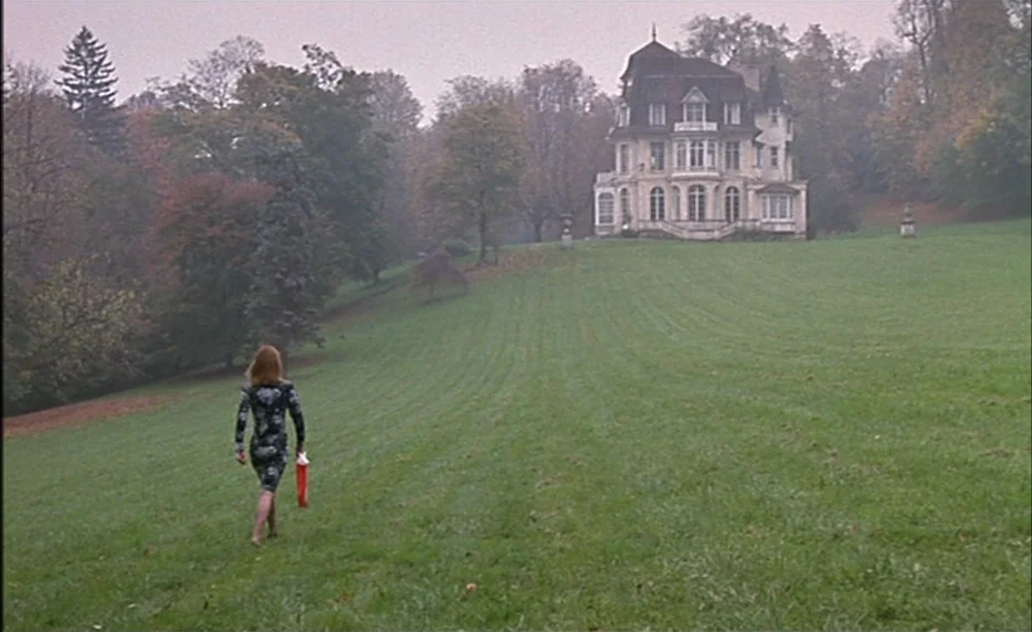 I Want To Go Home (1989) movie still: wide — A large multi-story manor sits atop a grassy hill surrounded by autumnal trees. A woman in a patterned…