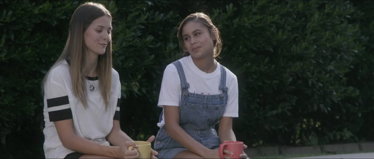 If You're Gone (2019) movie still: medium — Two women sit outdoors in front of a dense green hedge. They hold colorful mugs and look toward one…