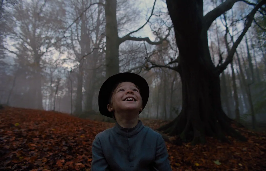 Gretel & Hansel (2020) movie still: medium — An autumnal forest floor covered in fallen brown leaves is shrouded in thick fog. A young boy in a hat…