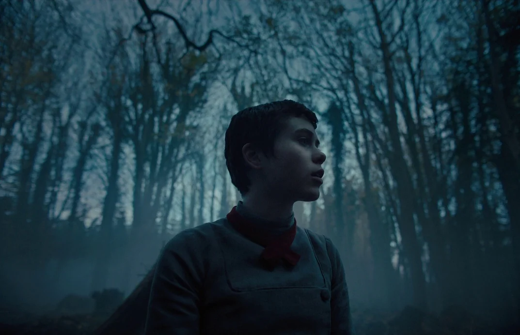 Gretel & Hansel (2020) movie still: medium — A dark, dense forest filled with tall, bare trees and swirling ground-level fog. A teenage girl with…