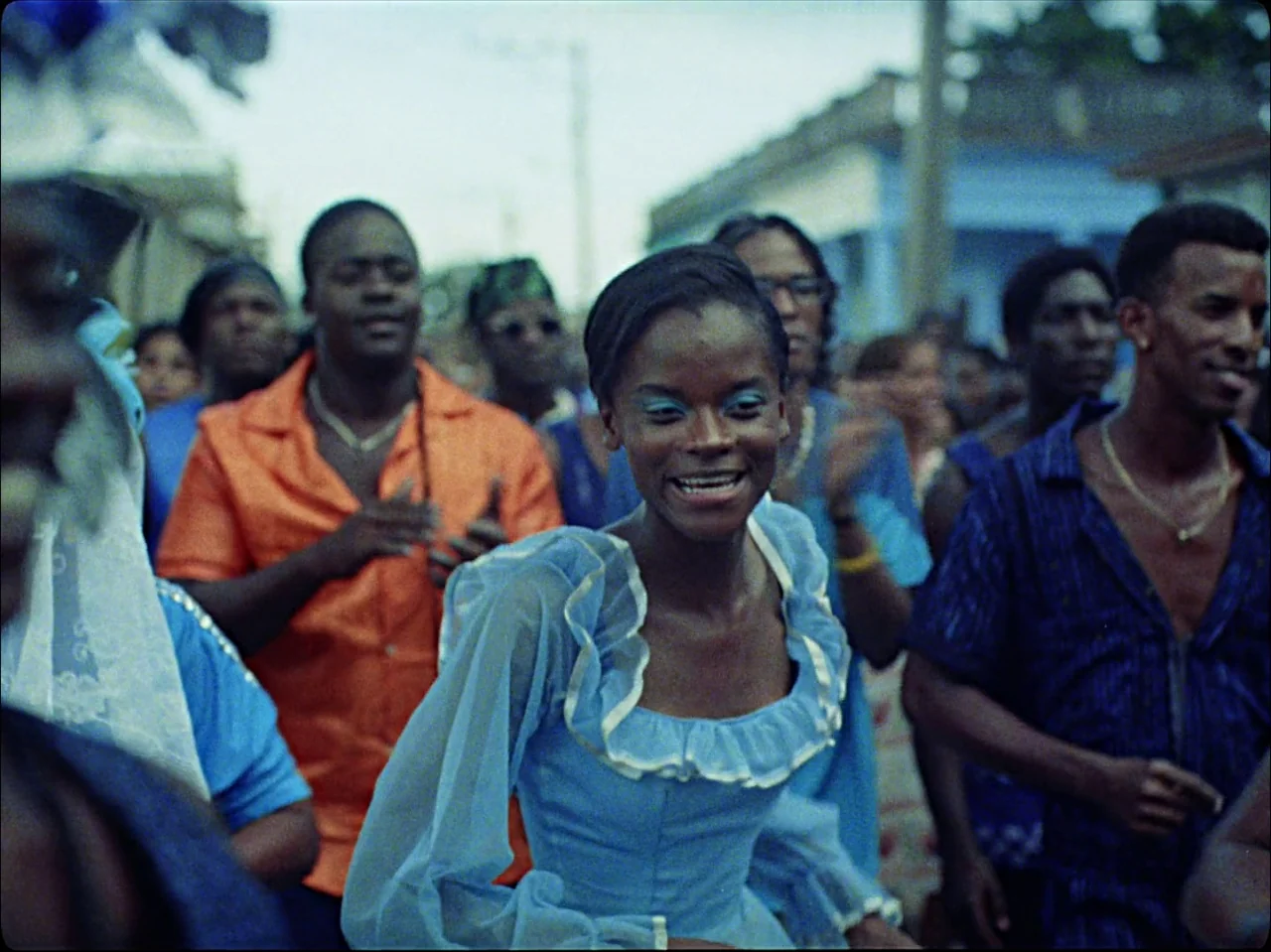 Guava Island (2019) movie still: medium — A crowd of people celebrate in an outdoor street setting during the day. A young woman in a blue dress…