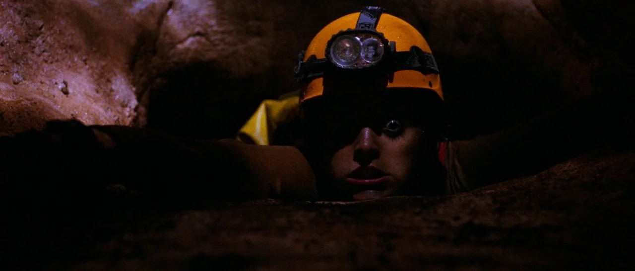 The Descent (2005) movie still: closeup — A narrow, dark limestone crawlspace features rough rock walls and limited visibility. A person crawls…