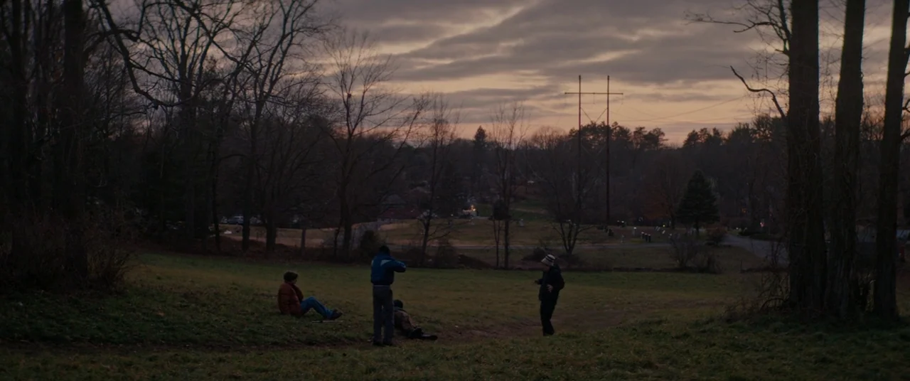 Super Dark Times (2017) movie still: wide — An open grassy field framed by bare trees under a dim, cloudy dusk sky. Four people stand or sit on the…