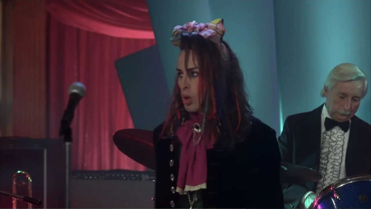 The Wedding Singer (1998) movie still: medium — A dimly lit club stage with red curtains and teal structural pillars. A lead singer with colorful…