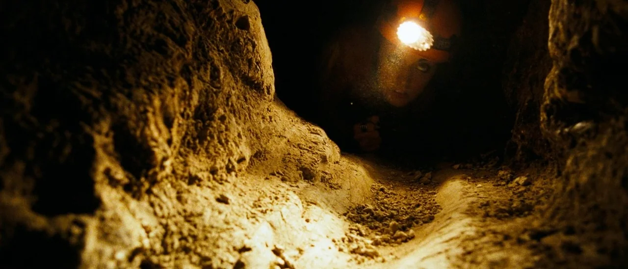 The Descent (2005) movie still: medium — A narrow, dirt-floored cave passage is framed by rough, irregular rock walls. A woman crawls through the…