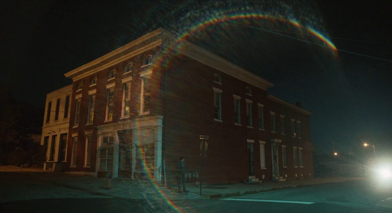 The Art of Self Defence (2019) movie still: wide — A dark brick building stands on a quiet street corner at night. A bright headlight beam cuts…