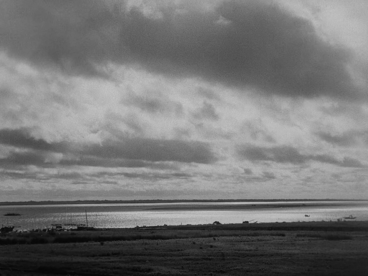 The Music Room (1958) movie still: establishing — A wide river stretches to the horizon beneath a sky filled with heavy clouds. A grassy bank…