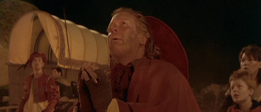 Flesh+Blood (1985) movie still: medium — A group of people gathers in a dark encampment at night near a large wagon. A man in a red cloak gazes…