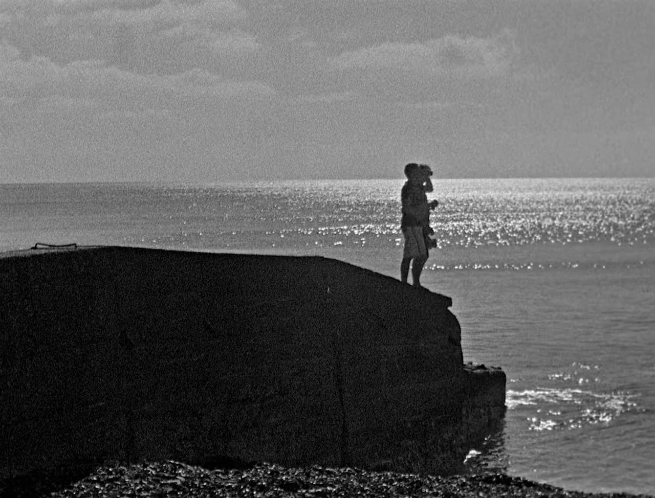 Bait (2019) movie still: wide — A man stands on a weathered concrete seawall overlooking a calm, vast ocean. The bright sun glints off the water…