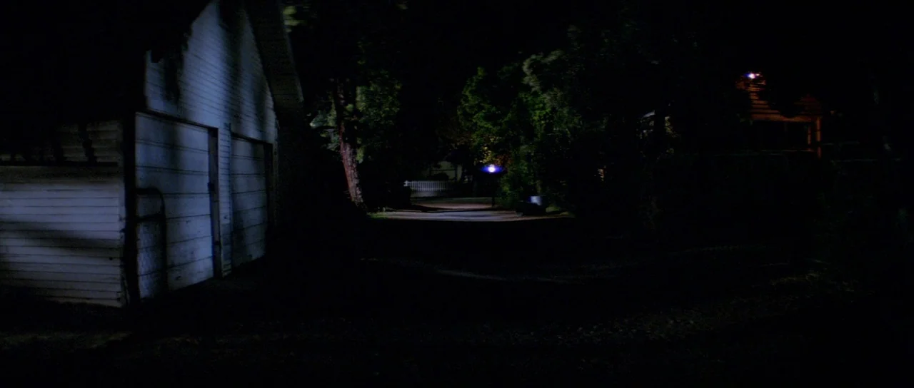 Halloween II (1981) movie still: establishing — A quiet residential alley at night features a white wooden garage in the foreground. A distant…