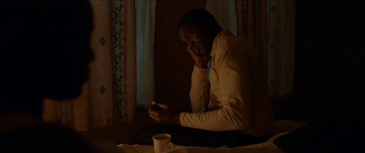 His House (2020) movie still: medium — A dimly lit room with patterned curtains visible in the background. A man sits on the side of a bed holding a…