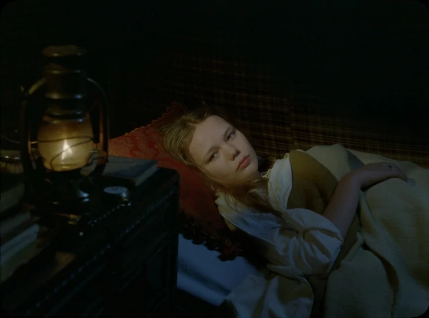 Jauja (2014) movie still: medium — A dark bedroom features a wooden bed frame covered with a plaid headboard cloth. A teenage girl with blonde hair…