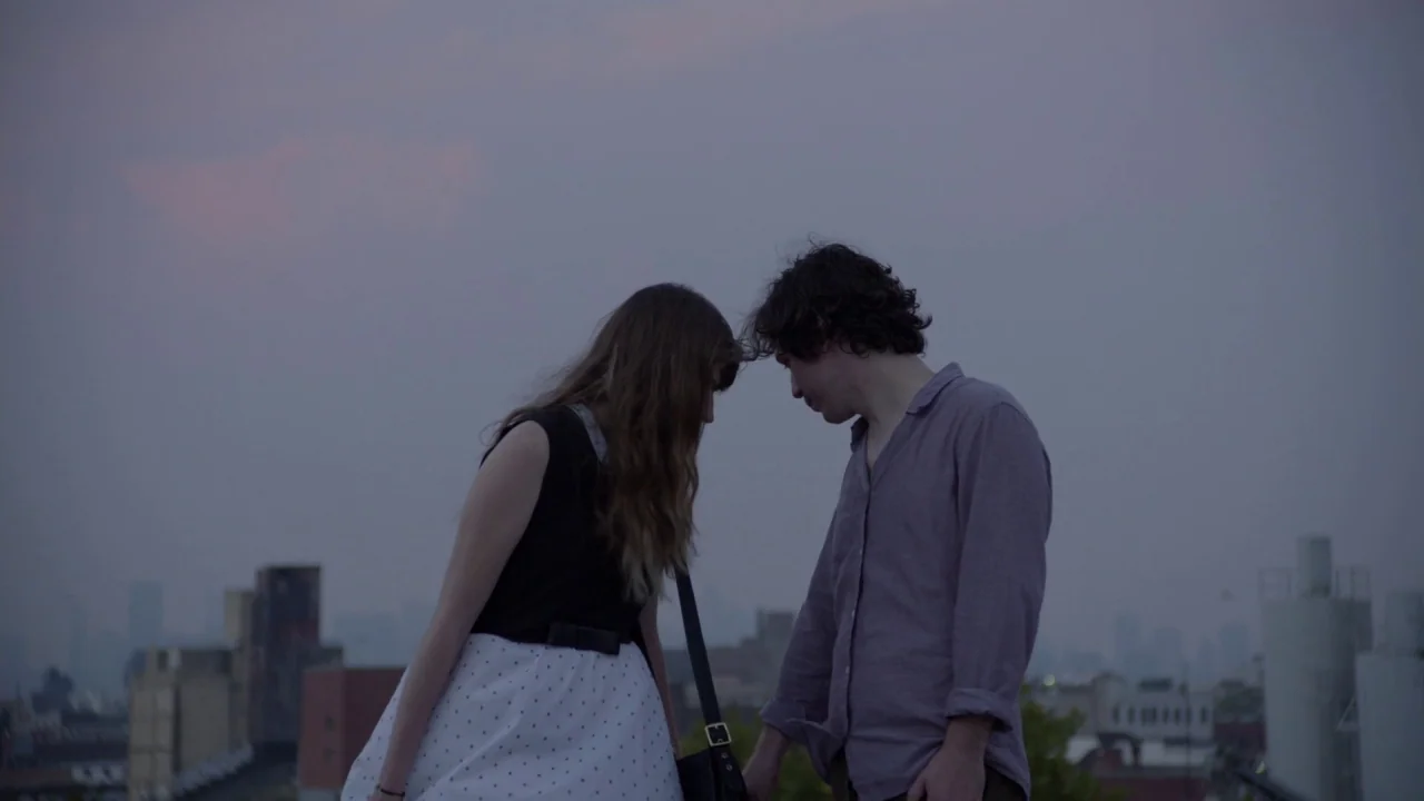 The Heart Machine (2014) movie still: medium — A rooftop terrace at dusk overlooks a blurry city skyline under a grey sky. A young man and woman…