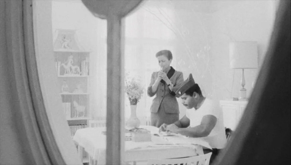 Veronika Voss (1982) movie still: medium — A light-filled 1950s dining room features a white table and decorative shelving units. A man in a…