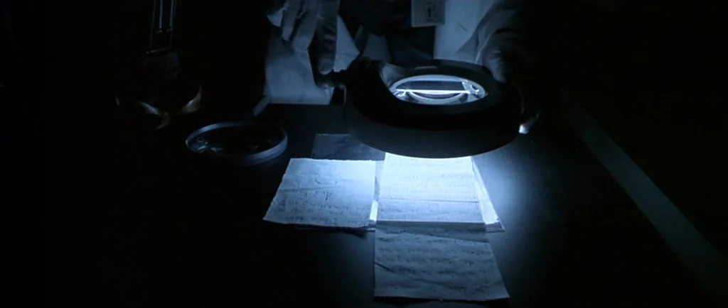 Manhunter (1986) movie still: closeup — A dark workspace features a white laboratory coat and documents placed on a desk. A glowing magnifying glass…