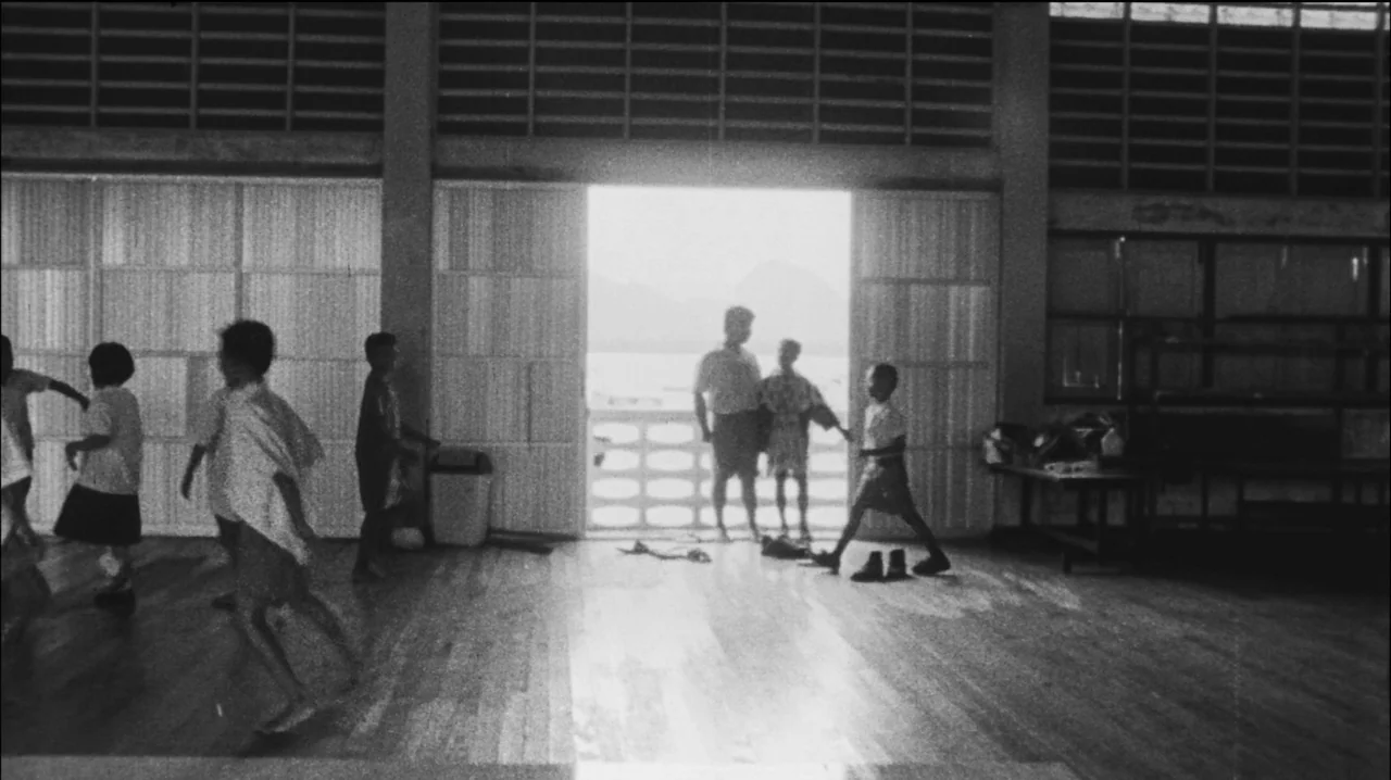 Mysterious Object at Noon (2000) movie still: wide — A spacious wooden-floored room features louvered walls and a large open doorway leading…