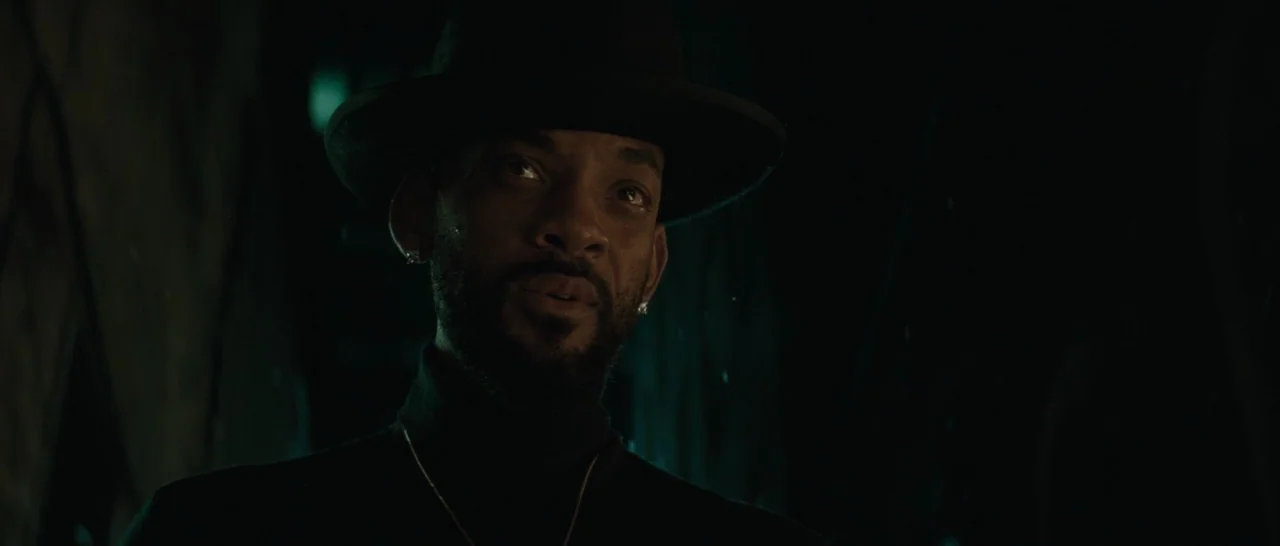 Suicide Squad (2016) movie still: closeup — A man stands in an dark outdoor space with indistinct shadowy forms behind him. The man looks toward the…