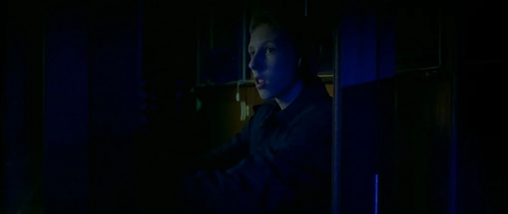 I Want You (1998) movie still: medium — A dimly lit, narrow interior space with dark walls and minimal environmental detail. A young man wearing…