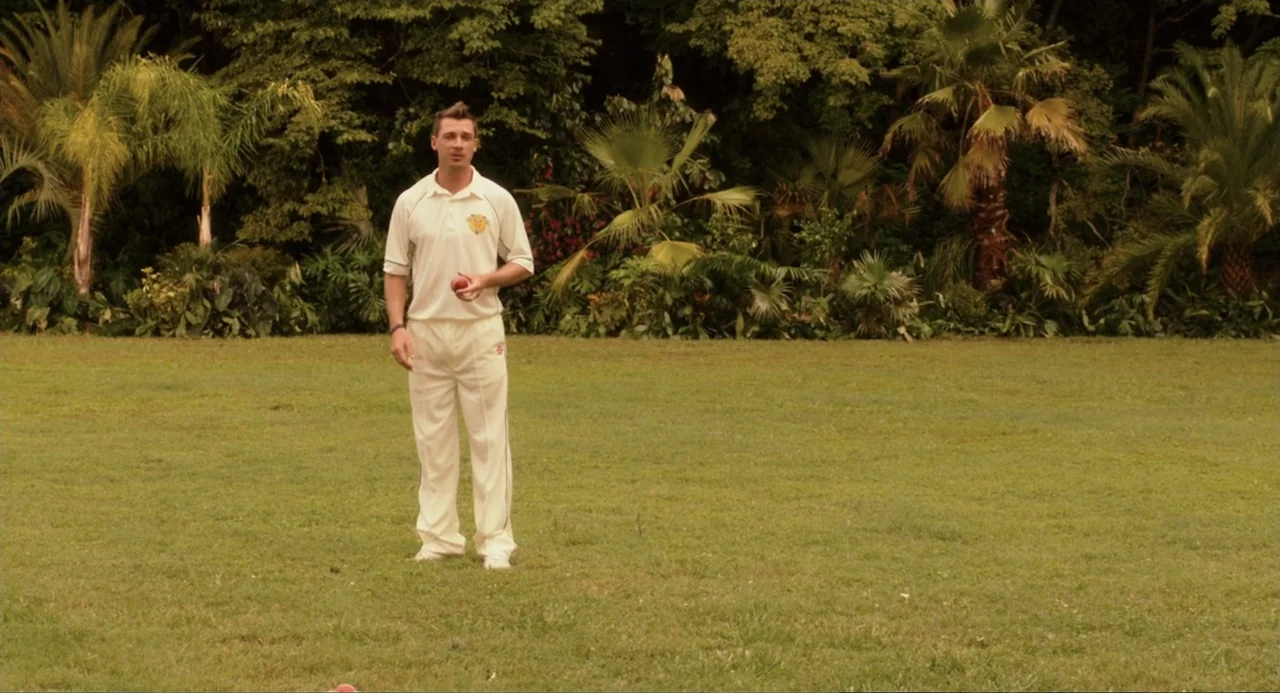 Blended (2014) movie still: fullbody — A man stands in the center of a manicured grassy sports field bordered by trees. He wears white cricket…