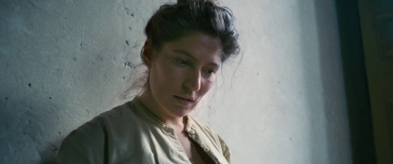 At Eternity’s Gate (2018) movie still: closeup — A woman stands indoors against a rough, light-colored concrete or stone wall. She gazes downward…