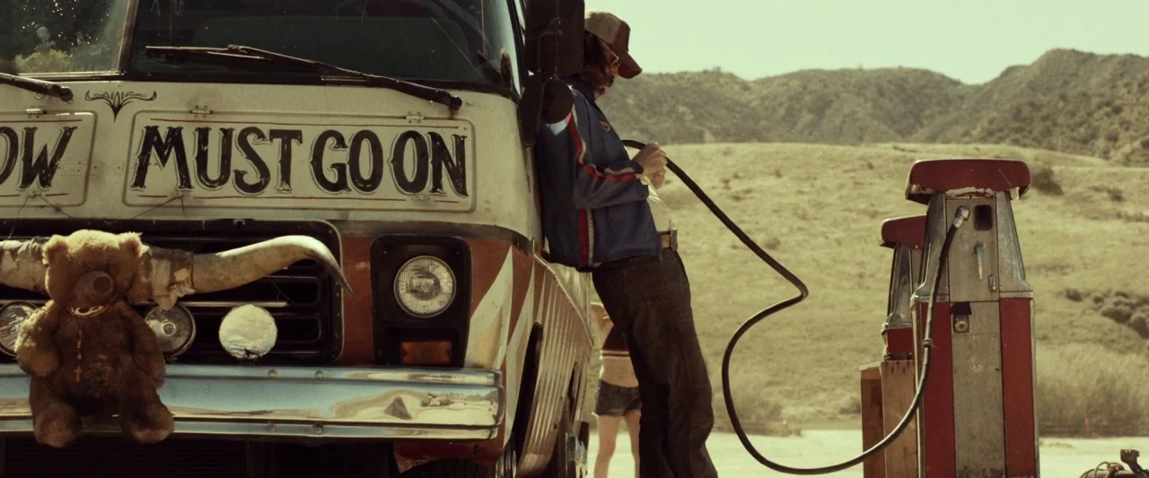 31 (2016) movie still: medium — A vintage bus with a teddy bear ornament is parked at an arid desert station. A man with a beard and baseball cap…