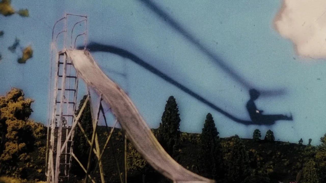 Once Within A Time (2022) movie still: wide — A metal slide stands on a grassy hill with tall cypress trees in distance. A prominent dark shadow of…