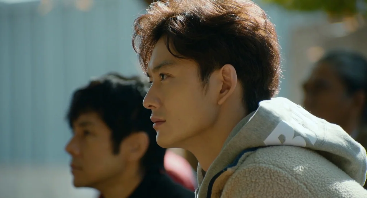 Drive My Car (2021) movie still: medium — Two men sit outside in a bright, outdoor setting during the day. They face the same direction while…