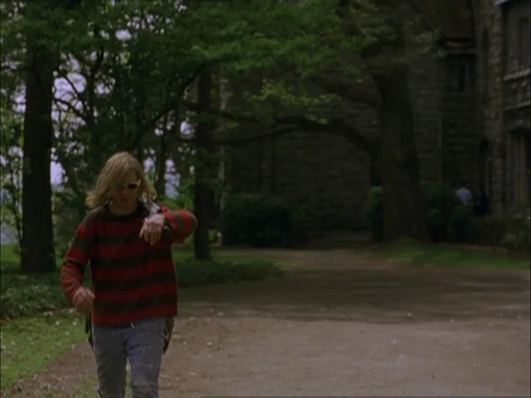 Last Days (2005) movie still: fullbody — An outdoor campus driveway is lined with mature trees and a grey stone building. A young man wearing a…