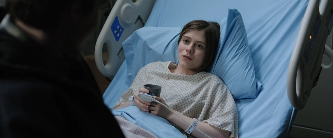 Trap House (2025) movie still: medium — A hospital bed with light blue linens occupies the center of a brightly lit room. A young woman wearing a…