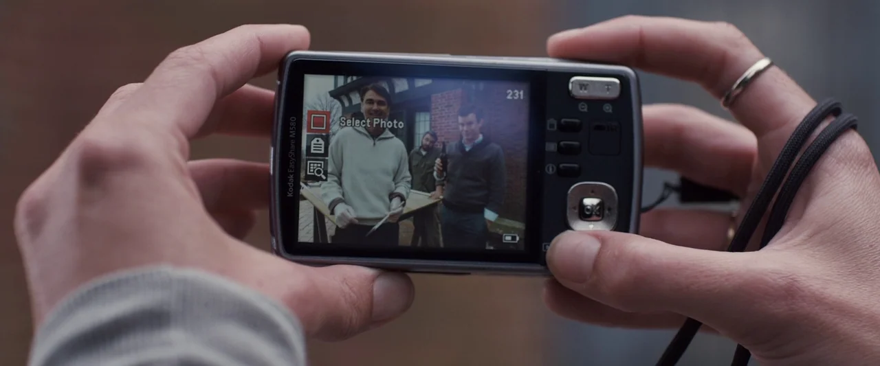 You’re Next (2013) movie still: closeup — A pair of hands holds a compact digital camera displaying a photograph. The screen shows three men…