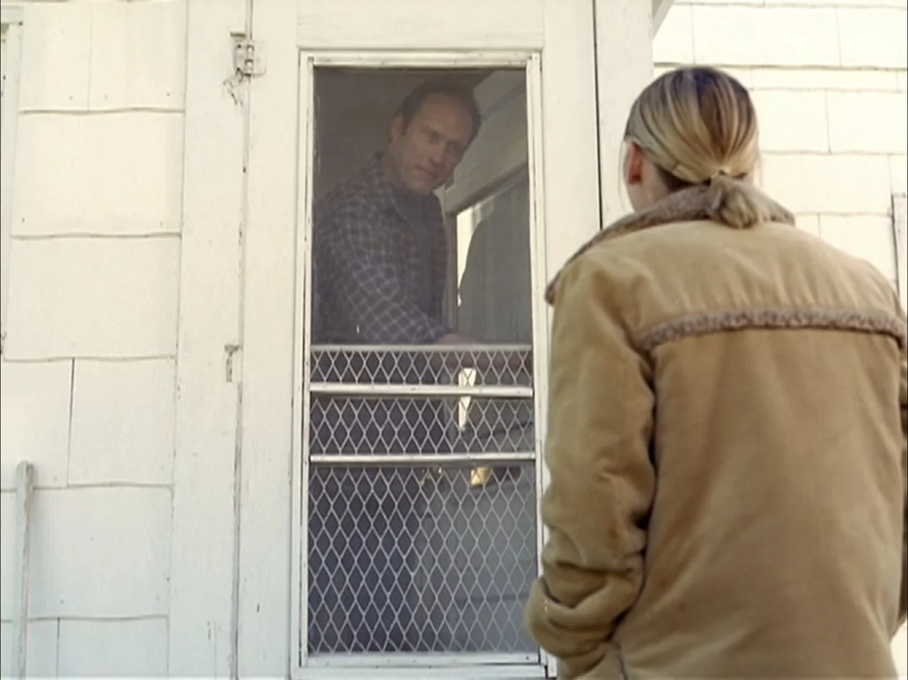 The Laramie Project (2020) movie still: medium — A woman stands before a white-painted wooden house with a screen door. A man visible behind the…