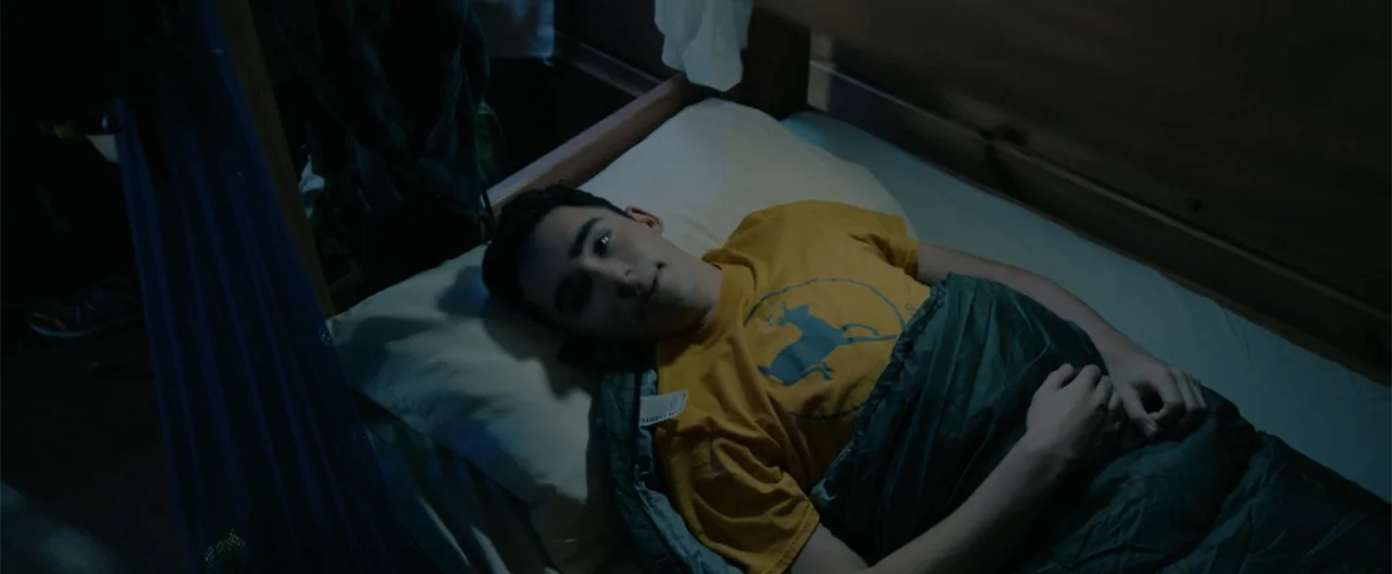 Camp Hideout (2023) movie still: medium — A dark, cramped interior of a wooden bunk bed frame at night. A young male lies on his back in a sleeping…