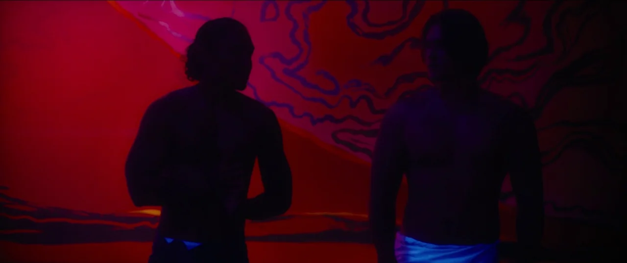 Pacifiction (2022) movie still: medium — A dimly lit room with a vibrant red wall featuring abstract purple graphic patterns. Two shirtless men with…