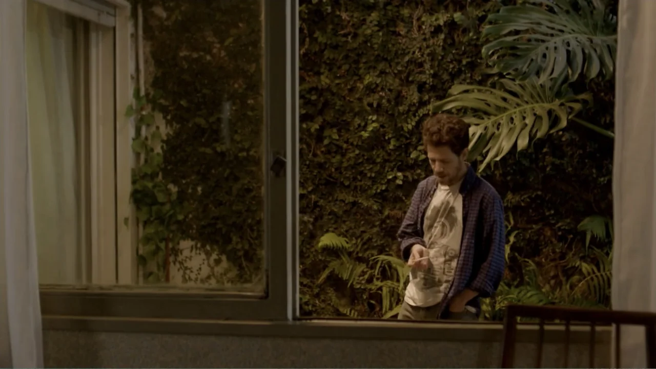 Heaven Sent (1994) movie still: medium — A man stands indoors next to a large window revealing a dark ivy-covered wall. He holds a small slender…