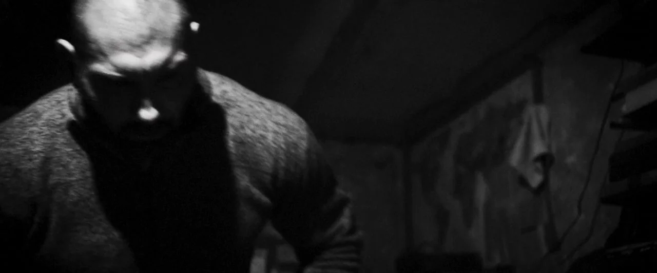 Bushwick (2017) movie still: medium — A dimly lit, shadowed room features a man standing in the foreground. He leans downward, his upper body…