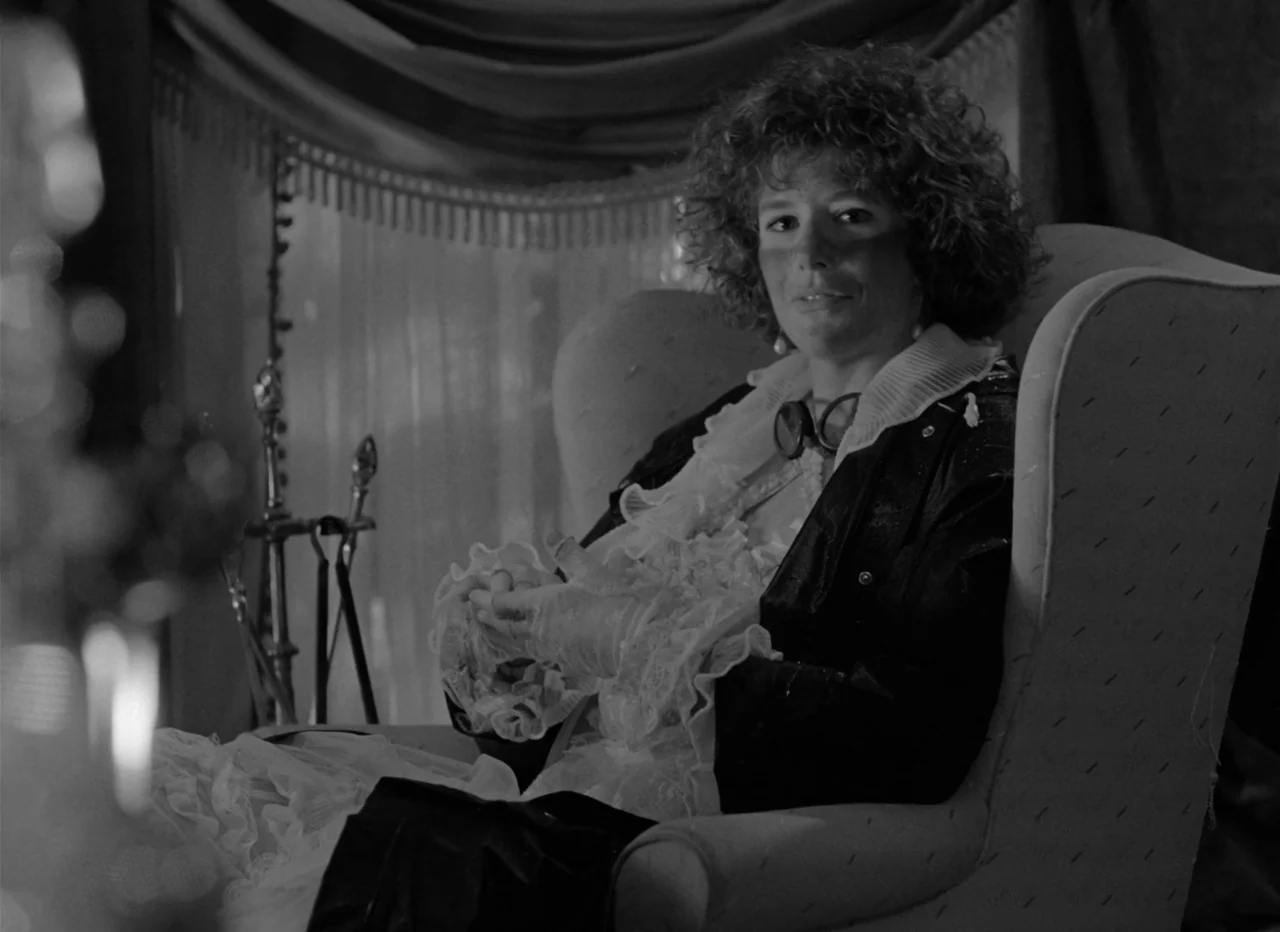 Singapore Sling (1990) movie still: medium — A person sits in a high-backed armchair in a room with sheer curtains. The subject wears a leather…