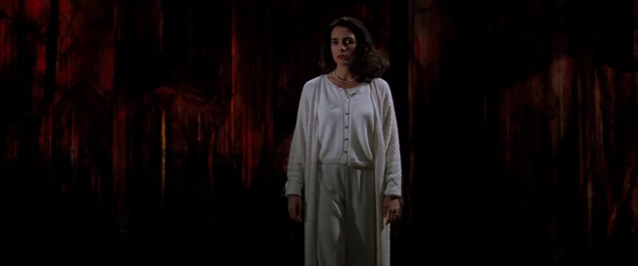 In The Mouth of Madness (1994) movie still: medium — A woman stands centered in front of a dark red textured backdrop. She is illuminated by a…