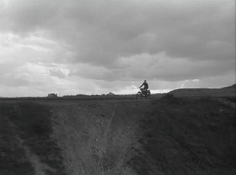 Head Against The Wall (1959) movie still: wide — A vast, open countryside landscape under a heavy, overcast cloudy sky. A solitary person rides a…