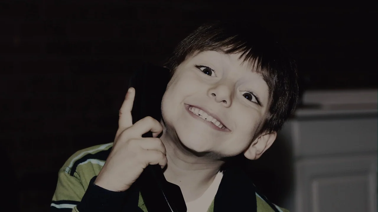 Saw III (2006) movie still: closeup — A dark indoor space with a dimly lit wall behind the subject. A young boy smiles directly into the camera…