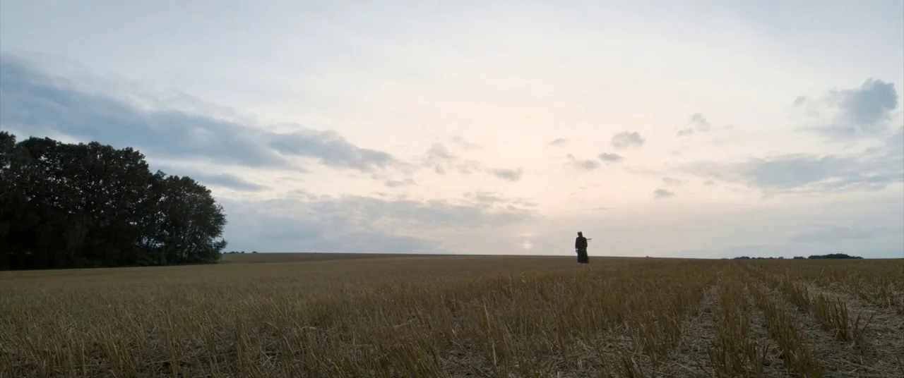 A Hidden Life (2019) movie still: wide — A vast harvested wheat field stretches toward a tree line under a cloudy sky. A solitary figure stands at a…