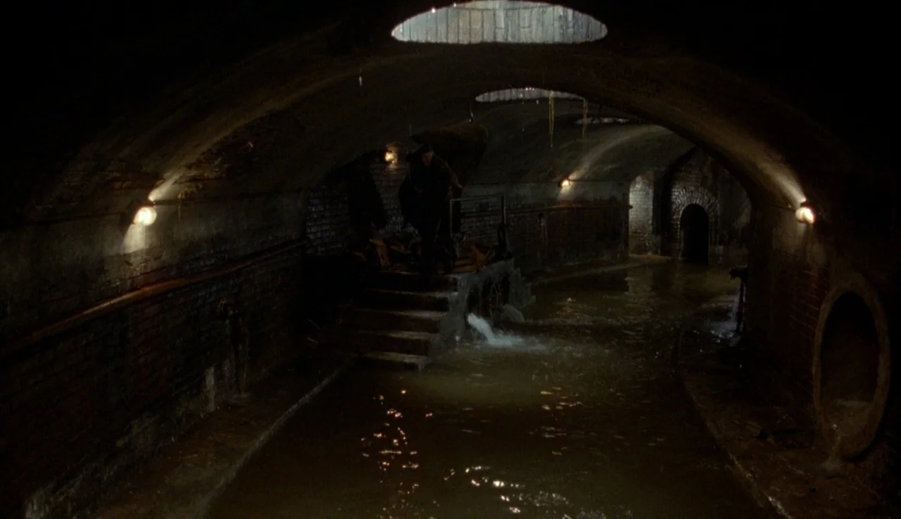 The Rainbow Thief (1990) movie still: wide — A dark subterranean brick sewer tunnel with arched ceilings and murky standing water. Light streams…
