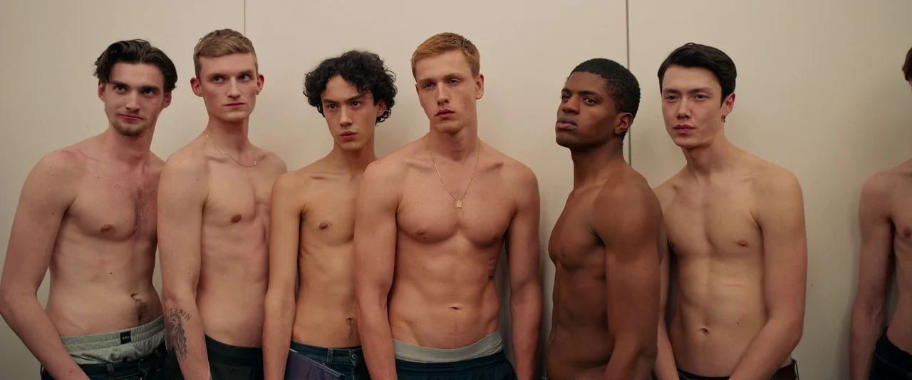 Triangle of Sadness (2022) movie still: medium — A group of shirtless men stand in a single line against a plain wall. They face the camera with…