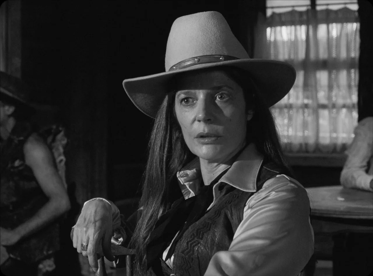 Eureka (2024) movie still: medium — A rustic saloon interior features a woman sitting at a wooden table. She wears a cowboy hat and stares directly…