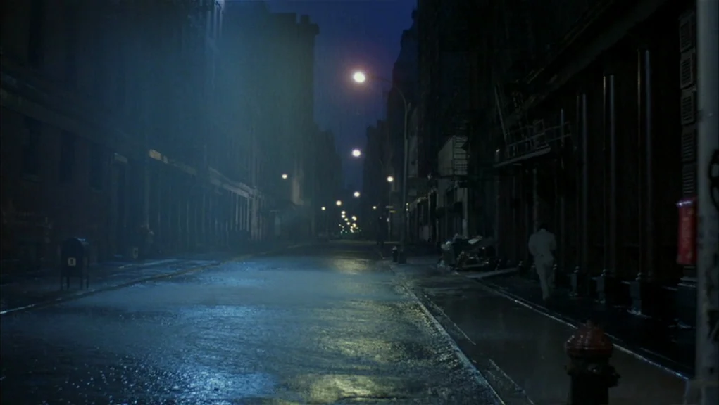 After Hours (1985) movie still: wide — An empty urban thoroughfare is slick with rain under the glow of streetlights. A single figure in…