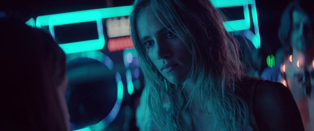 The Bad Batch (2016) movie still: closeup — A dimly lit nightclub with vibrant cyan neon signage glowing in the background. A young woman with…