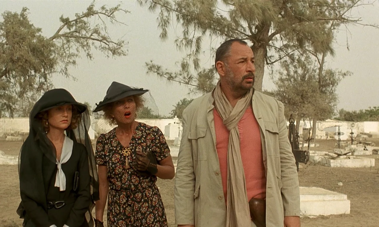 Coup De Torchon (1981) movie still: fullbody — An outdoor cemetery with dry dirt, scattered white headstones, and dry trees. Three adults stand…