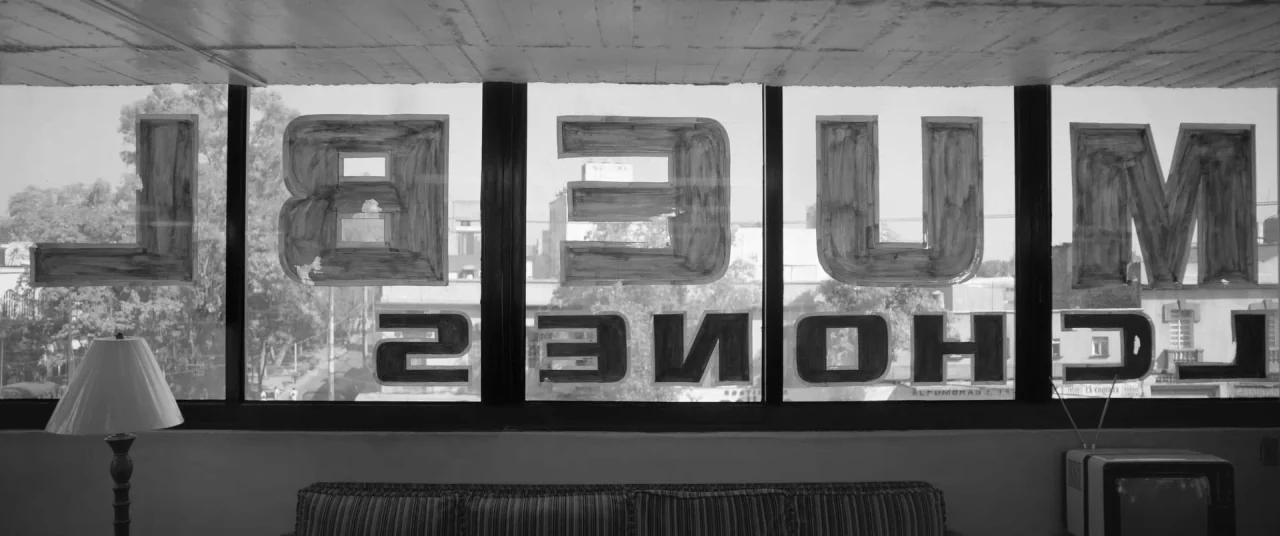 Roma (2018) movie still: wide — A room interior features a long horizontal window looking out over a city street. Large painted letters are applied…