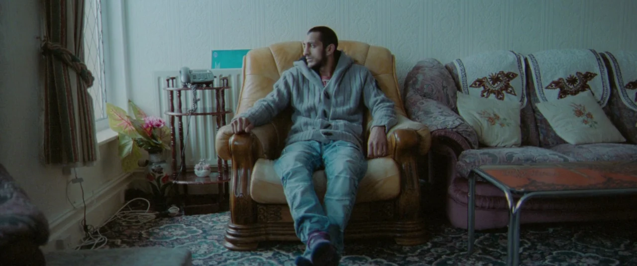 Catch Me Daddy (2014) movie still: medium — A dated interior space features floral patterned carpeting, a brown leather armchair, and wallpaper. A…