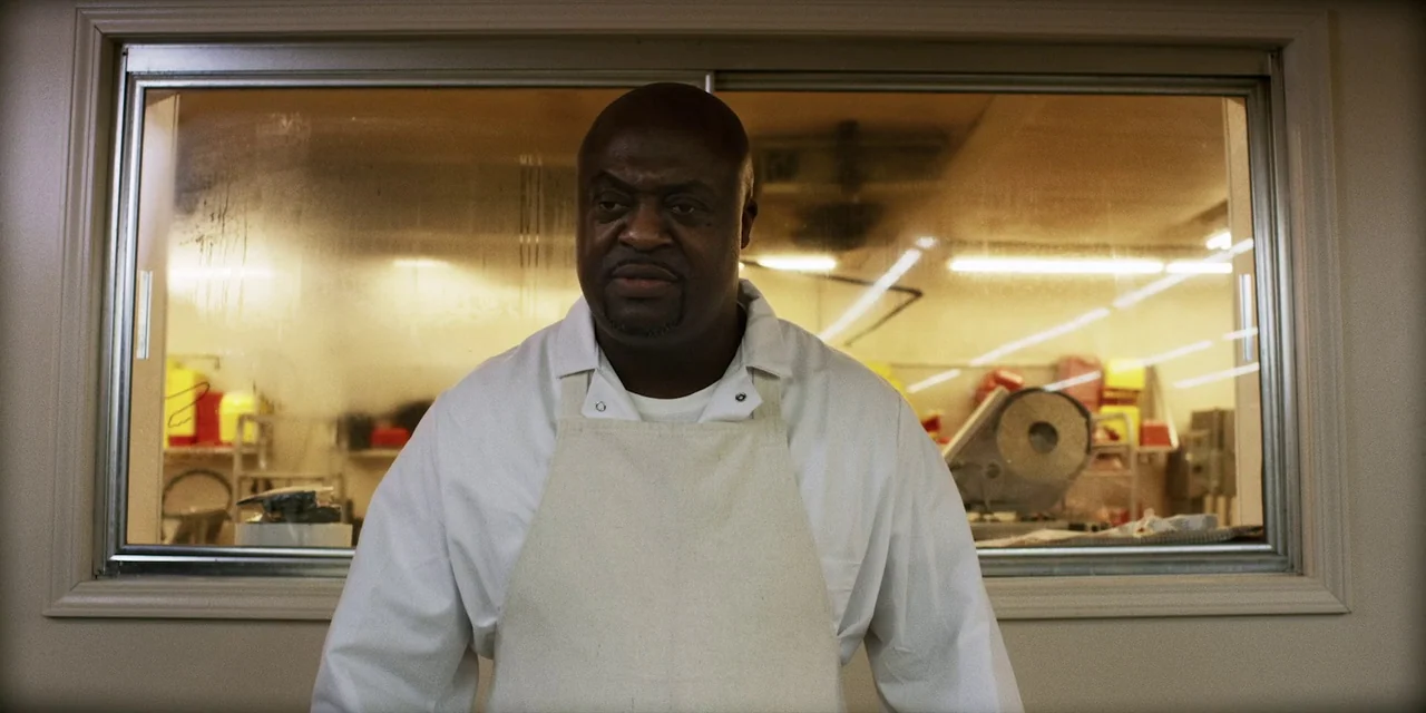 Jakob’s Wife (2021) movie still: medium — A man stands before a large glass window separating him from a commercial food prep area. Behind the glass…
